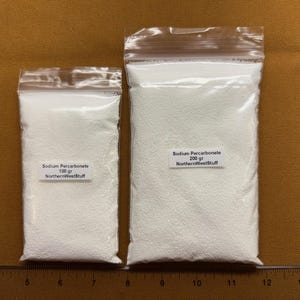 May include: Two clear plastic bags filled with white powder, labeled "Sodium Percarbonate." One bag is labeled 100 gr, the other 200 gr. The bags are on a brown surface, with a ruler in the foreground. The text "NorthernWestStuff" is also visible.