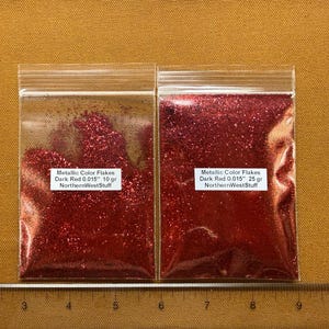 May include: Two clear plastic bags filled with dark red metallic glitter flakes. The bags have white labels with text that reads "Metallic Color Flakes Dark Red 0.015"" and "NorthernWestStuff". A ruler is at the bottom.