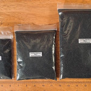 May include: Three clear plastic bags filled with black coal slag. The bags are labeled with the product name, mesh size, and weight in grams. A wooden ruler is at the bottom of the image.