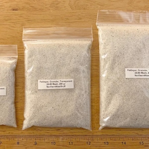 May include: Three clear plastic bags filled with granular feldspar. Each bag has a label with product details. The bags contain 100g, 200g, and 400g of feldspar, respectively. The bags are on a wooden surface.
