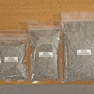 May include: Three clear plastic bags filled with small, gray gravel-like material. Each bag has a white label with text indicating the contents are Bauxite, with weights of 100, 200, and 400 grams. A metal ruler is at the bottom.