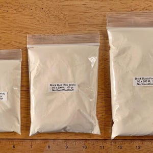 May include: Three clear plastic bags filled with light-colored brick dust, labeled with product details. The bags are of varying sizes, with the largest containing 200 grams. The bags are arranged on a wooden surface.