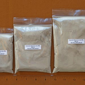 May include: Three clear plastic bags of cement mix, labeled "Cement All Concrete Mix" with weights of 100g, 200g, and 400g. The bags are sealed and arranged on a brown surface next to a ruler.