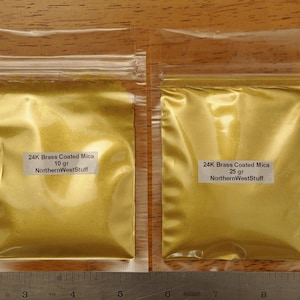 May include: Two clear plastic bags filled with gold-colored powder, labeled "24K Brass Coated Mica." One bag is labeled 10 gr, the other 25 gr. The text "NorthernWestStuff" is also visible on the labels. A metal ruler is at the bottom.