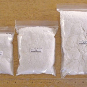 May include: Three clear plastic bags filled with white paper pulp. Each bag has a label with text. The bags are of varying sizes, with the largest containing 100 grams of pulp. A wooden ruler is at the bottom.