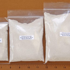 May include: Three clear plastic bags filled with small, off-white aragonite beads. Each bag has a label indicating the contents: 100 grams, 200 grams, and 400 grams. The bags are arranged in a row, with a ruler at the bottom.