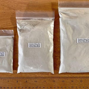 May include: Three clear plastic bags filled with light green ceramic glaze powder. Each bag has a white label with text indicating the weight: 50 grams, 200 grams, and 400 grams. The bags are on a wooden surface.