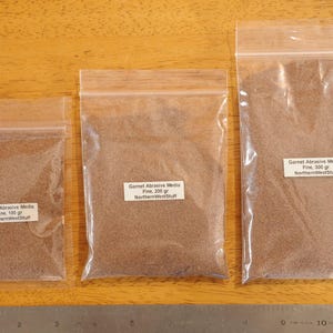 May include: Three clear plastic bags filled with fine, reddish-brown garnet abrasive media. Each bag has a white label with text indicating the weight: 100 gr, 200 gr, and 300 gr. A metal ruler is at the bottom.