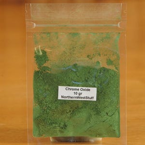 May include: A clear plastic bag filled with vibrant green Chrome Oxide powder. A white label displays the text "Chrome Oxide 10 gr NorthernWestStuff". The bag is sealed and set against a blurred wooden background.