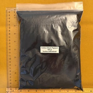 May include: A clear plastic resealable bag filled with fine carbon black pigment powder. A white label on the front reads Carbon Black Pigment 100 gr NorthernWestStuff. A clear plastic ruler is placed along the side and bottom for scale.