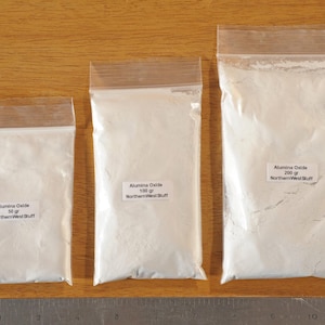 May include: Three clear plastic bags filled with white powder, labeled "Alumina Oxide" with weights of 50g, 100g, and 200g. The bags are sealed and placed on a wooden surface. The text "NorthernWestStuff" is also visible.