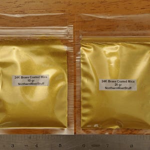 Puede incluir: Dos bolsas de pl&aacute;stico transparente llenas de polvo dorado, etiquetadas como "24K Brass Coated Mica". Una bolsa est&aacute; etiquetada con 10 g, la otra con 25 g. El texto "NorthernWestStuff" tambi&eacute;n es visible en las etiquetas. Una regla en la parte inferior.