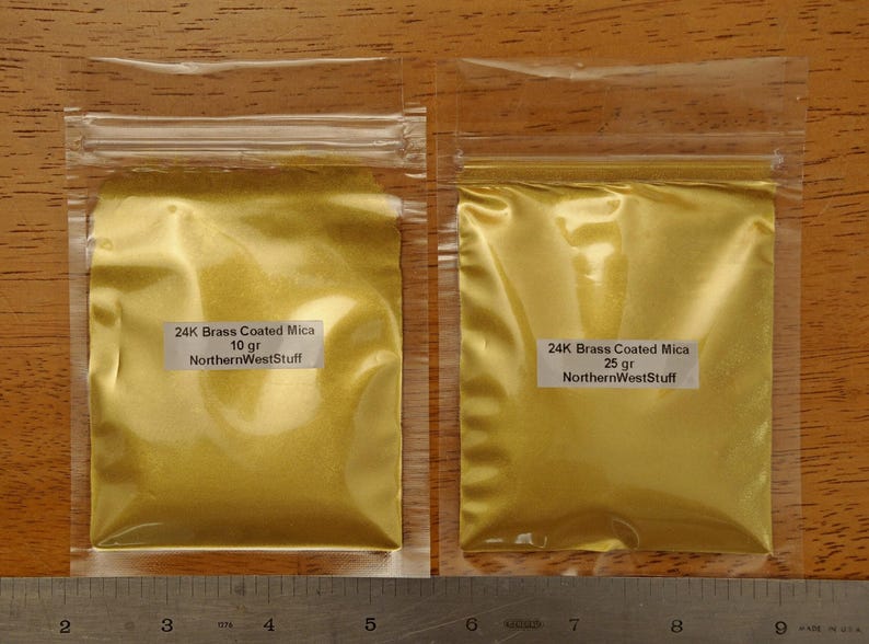 Puede incluir: Dos bolsas de pl&aacute;stico transparente llenas de polvo dorado, etiquetadas como "24K Brass Coated Mica". Una bolsa est&aacute; etiquetada con 10 g, la otra con 25 g. El texto "NorthernWestStuff" tambi&eacute;n es visible en las etiquetas. Una regla en la parte inferior.