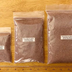 May include: Three clear plastic bags filled with garnet abrasive media, each with a white label. The bags contain 100 grams, 400 grams, and 800 grams of the abrasive material. The bags are on a wooden surface.