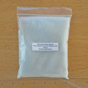 Puede incluir: Una bolsa de plástico transparente llena de cuentas de vidrio reflectantes. La etiqueta blanca dice "Reflective Glass Beads", "Very Coarse (0.74 mm Median Dia)", "400 gr", y "NorthernWestStuff". Las cuentas son claras y gruesas.
