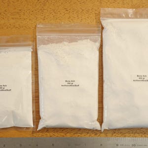 May include: Three clear plastic bags filled with white powder, labeled "Bone Ash" with weights of 100 grams, 200 grams, and 400 grams. A metal ruler is at the bottom of the image, with measurements in inches.