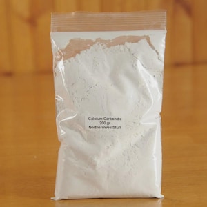 May include: A clear plastic bag filled with white powder, labeled "Calcium Carbonate 200 gr NorthernWestStuff." The bag is sealed at the top and sits on a light brown wooden surface. The powder appears fine and uniform.