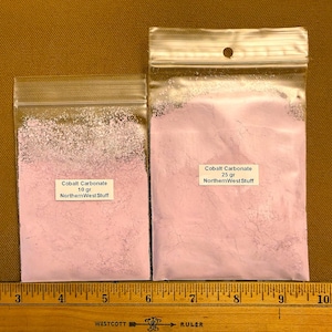 May include: Two clear plastic resealable bags containing light purple cobalt carbonate powder. The smaller bag is labeled 10 gr and the larger bag is labeled 25 gr, both with the text NorthernWest Stuff, positioned above a yellow ruler.