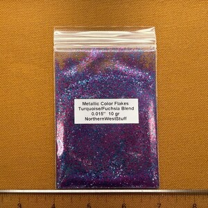 May include: A clear plastic bag filled with metallic color flakes in a turquoise and fuchsia blend. The bag contains 10 grams of flakes, with a size of 0.015 inches. The label reads "Metallic Color Flakes Turquoise/Fuchsia Blend 0.015" 10 gr NorthernWestStuff."