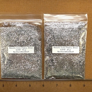 May include: Two clear plastic bags filled with silver metallic flakes. Each bag has a white label with black text that reads "Metallic Color Flakes, Silver 0.015" 10 gr" and "Metallic Color Flakes, Silver 0.015" 25 gr". The brand name "NorthernWestStuff" is also printed on the labels.