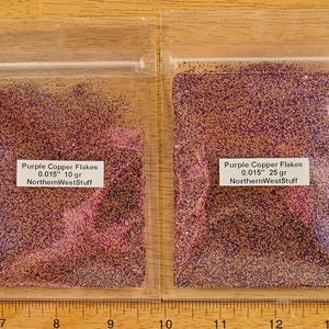 May include: Two clear plastic bags filled with purple and copper-colored glitter flakes. The bags have white labels with the text "Purple Copper Flakes 0.015" 10 gr NorthernWestStuff" and "Purple Copper Flakes 0.015" 25 gr NorthernWestStuff".