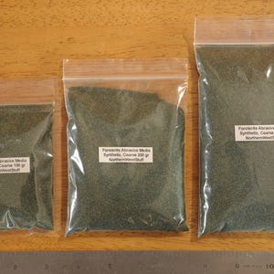 May include: Three clear plastic bags filled with green abrasive media, labeled "Forsterite Abrasive Media Synthetic, Coarse" with varying weights of 100g, 200g, and 300g. A metal ruler is at the bottom.