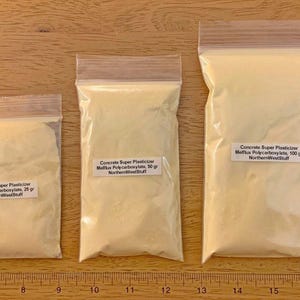 May include: Three clear plastic bags containing a light yellow powder, labeled "Concrete Super Plasticizer." The bags are labeled with the weight of the contents: 25 grams, 50 grams, and 100 grams. The bags are on a wooden surface.