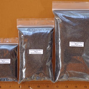 May include: Three clear plastic bags filled with dark gray coal slag. Each bag has a white label with black text indicating the product name, mesh size, and weight in grams. The bags are arranged from smallest to largest.