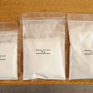 May include: Three clear plastic bags filled with white powder, labeled "Dolomite, 200 mesh." The bags contain 50 grams, 100 grams, and 200 grams of the powder. A ruler is placed below the bags.