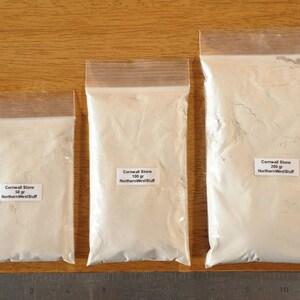 May include: Three clear plastic bags filled with a white powder, labeled "Cornwall Stone" with varying weights: 50 gr, 100 gr, and 200 gr. The bags are sealed and placed on a wooden surface next to a metal ruler.