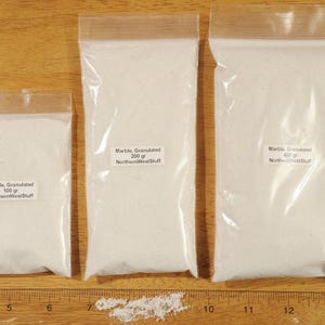 May include: Three clear plastic bags filled with white granulated marble. The bags are labeled with the weight of the marble in grams: 100 gr, 200 gr, and 400 gr. A wooden ruler is at the bottom of the image.