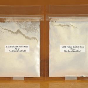 May include: Two clear plastic bags containing gold-tinted coated mica powder. The bag on the left is labeled "Gold Tinted Coated Mica 10 gr NorthernWestStuff." The bag on the right is labeled "Gold Tinted Coated Mica 25 gr NorthernWestStuff."