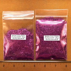 May include: Two clear plastic bags filled with fuchsia metallic color flakes. The bags have white labels with text that reads "Metallic Color Flakes Fuchsia 0.015" 10 gr NorthernWestStuff" and "Metallic Color Flakes Fuchsia 0.015" 25 gr NorthernWestStuff".