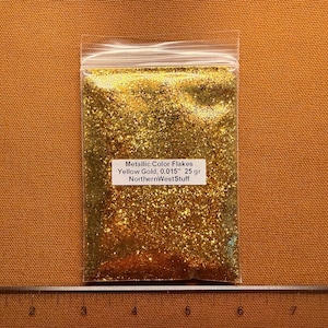 May include: A clear plastic bag filled with yellow gold metallic color flakes. The bag has a white label with the text "Metallic Color Flakes Yellow Gold, 0.015" 25 gr NorthernWestStuff". The bag is on a brown surface.