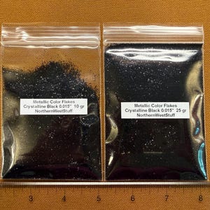 May include: Two clear plastic bags filled with black metallic flakes. The bags have labels that read "Metallic Color Flakes Crystalline Black 0.015" 10 gr NorthernWestStuff" and "25 gr NorthernWestStuff". A ruler is at the bottom.