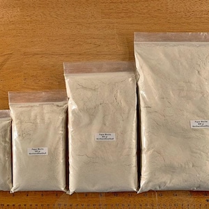 May include: Four clear plastic bags filled with a light beige powder, arranged in a row. Each bag has a label with text. The bags vary in size, with the largest bag containing the most powder. A wooden ruler is at the bottom.