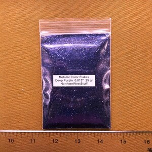 May include: A clear plastic bag filled with deep purple metallic color flakes. The bag has a white label with the text "Metallic Color Flakes Deep Purple 0.015" 25 gr NorthernWestStuff". A ruler is at the bottom.