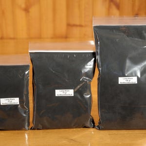 May include: Three sealed plastic bags filled with black powder, labeled "Lamp Black" with varying weights: 25g, 75g, and 100g. The bags are arranged on a wooden surface, with the labels indicating the product and the seller.