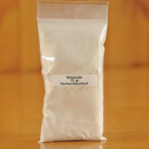 May include: A clear plastic bag filled with white powder, labeled "Metakaolin 75 gr NorthernWestStuff." The bag is sealed and set against a warm, wooden background.