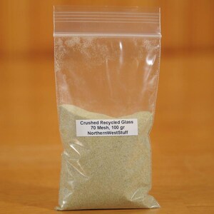 May include: A clear plastic bag filled with crushed, recycled glass. The bag has a white label with the text "Crushed Recycled Glass 70 Mesh, 100 gr NorthernWestStuff". The glass appears to be a light green color.
