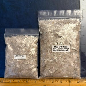 May include: Two clear plastic resealable bags filled with very coarse Muscovite Mica flakes. The smaller bag is labeled 50 gr and the larger bag is labeled 100 gr. A yellow ruler shows the width is approximately 10.5 inches and a ruler shows height.