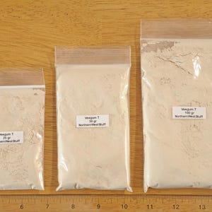 Puede incluir: Tres bolsas de plástico transparente llenas de un polvo beige claro, etiquetadas con pesos de 25 g, 50 g y 100 g. Las bolsas están selladas y colocadas sobre una superficie de madera. Las etiquetas dicen "Veegum T" y "Northern West Stuff."