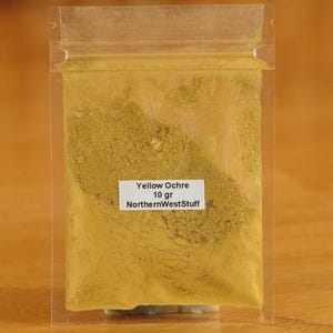 May include: A clear plastic bag filled with yellow ochre powder. A white label on the bag reads "Yellow Ochre 10 gr NorthernWestStuff". The powder has a fine, granular texture.