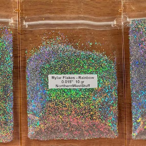 May include: Three clear plastic bags filled with iridescent rainbow mylar flakes. Each bag has a white label with text indicating the product name, flake size, and weight in grams. The text "NorthernWestStuff" is also visible.