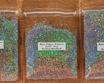 May include: Three clear plastic bags filled with iridescent rainbow mylar flakes. Each bag has a white label with text indicating the product name, flake size, and weight in grams. The text "NorthernWestStuff" is also visible.