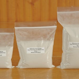 May include: Three clear plastic bags filled with white reflective glass beads. Each bag has a label with text that reads "Reflective Glass Beads Very Fine (0.02 mm Median Dia)" and "NorthernWestStuff". The bags contain 225g, 450g, and 900g of beads.