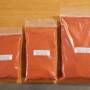 May include: Three clear plastic bags filled with red iron oxide powder. Each bag has a white label with text indicating the weight: 50g, 100g, and 200g. The bags are sealed and rest on a wooden surface.