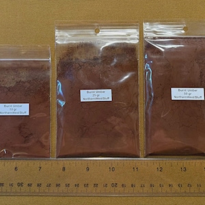 May include: Three clear plastic bags filled with brown powder labeled "Burnt Umber" in 10g, 25g, and 50g sizes. The bags are arranged in a row, with a ruler below them. The text "NorthernWestStuff" is also visible.