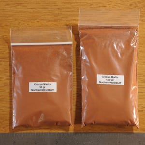May include: Two clear plastic bags filled with a reddish-brown powder, labeled "Crocus Martis." One bag is labeled 50 gr, the other 100 gr. The text "NorthernWestStuff" is also visible on the labels. The bags are on a wooden surface.