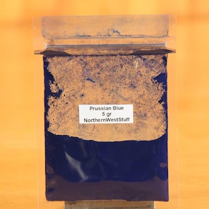 May include: A package of Prussian Blue pigment, with a label indicating 5 grams and the seller's name, NorthernWestStuff. The pigment is a deep blue color, with a lighter, textured area at the top. The package is sealed.